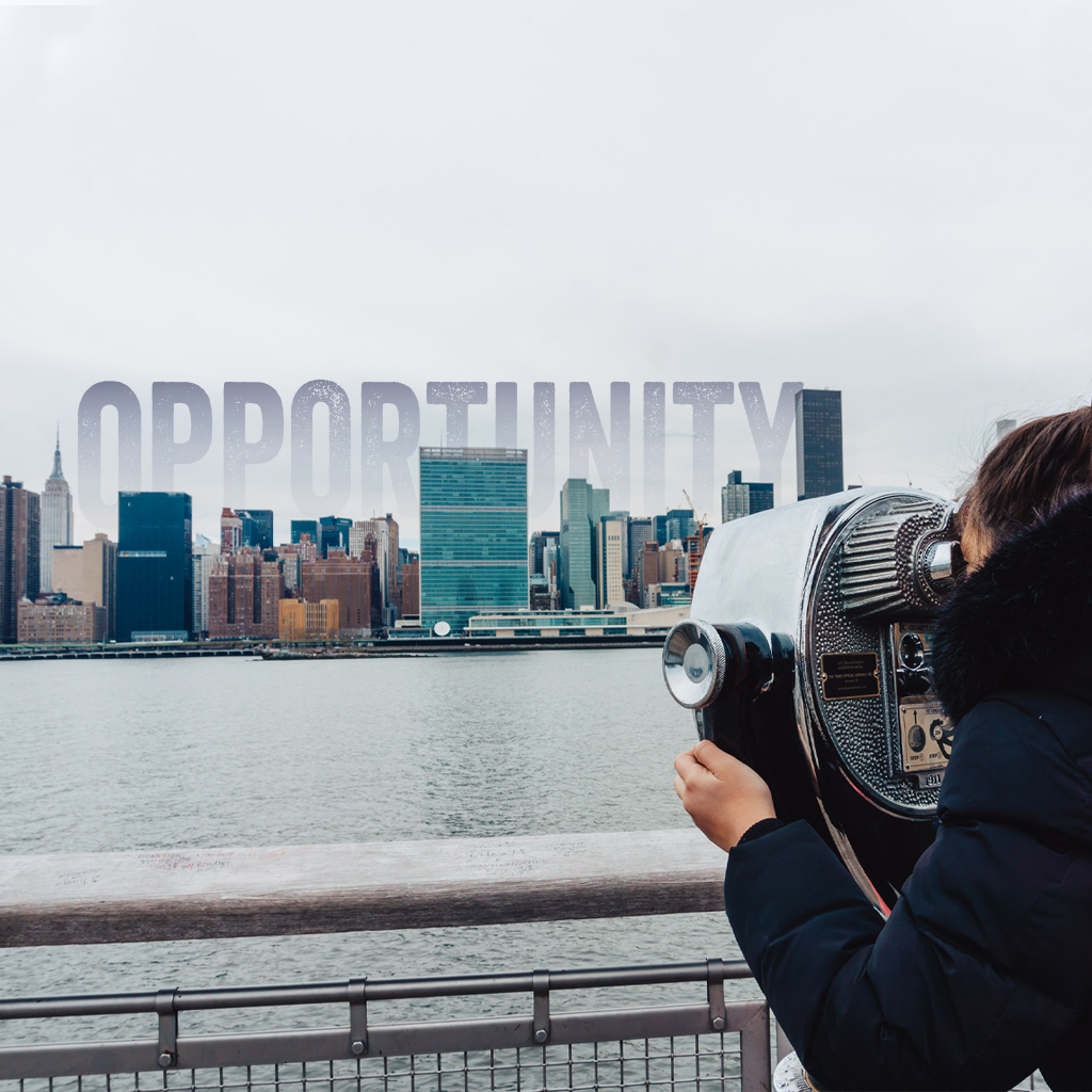 A person in a black fluffy coat with brown hair in a ponytail is looking through coin-operated binoculars across a body of water at a city skyline. Over the city the word "Opportunity" is superimposed on the sky.