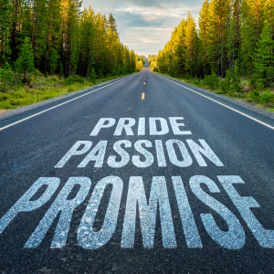 The words "pride, passion, promise" appear painted onto a rural 2-lane highway lines with evergreen trees