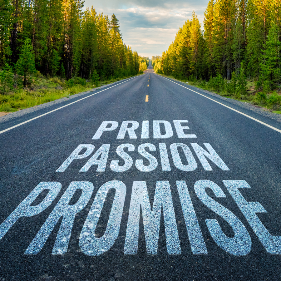 The words "pride, passion, promise" appear painted onto a rural 2-lane highway lines with evergreen trees